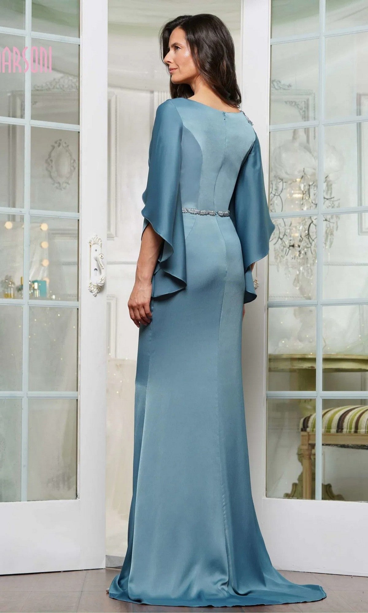 A woman stands indoors, facing away, in a floor-length teal Marsoni MV1348 formal dress with flutter sleeves, delicate beaded appliques, and a silver belt.